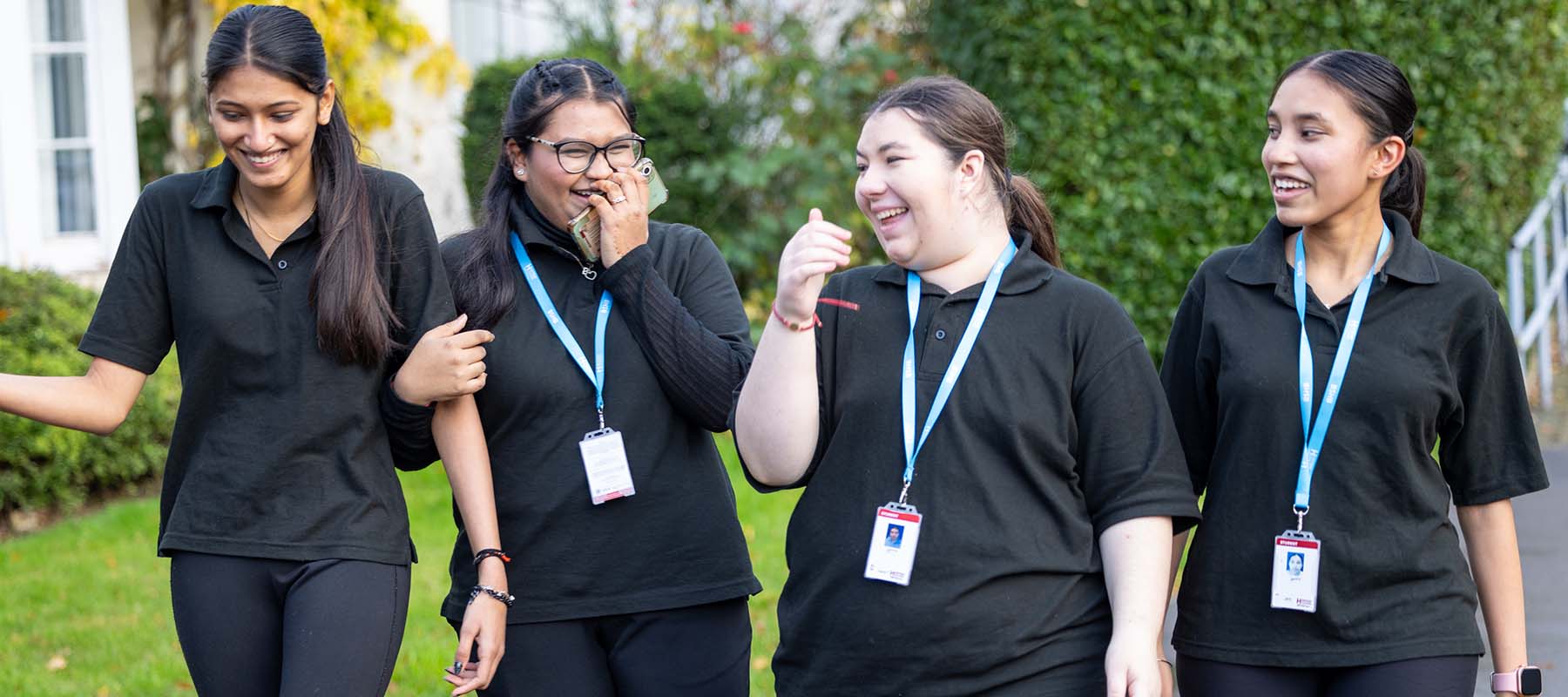 Four Harrow College students laughing and walking together. They are all wearing black with their lanyards around their necks.