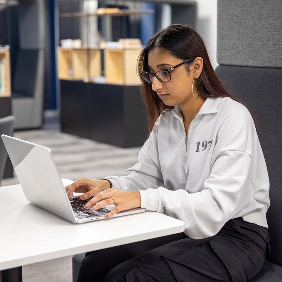 Uxbridge student sitting down at a desk and working on a laptop. She is wearing black glasses and a light coloured jumper.