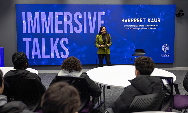 Former BBC The Apprentice winner and outstanding businesswoman Harpreet Kaur giving the first-ever Immersive Talk at Harrow College. Harpreet is standing in front of a blue screen, wearing a green jacket, with students sitting in the foreground.